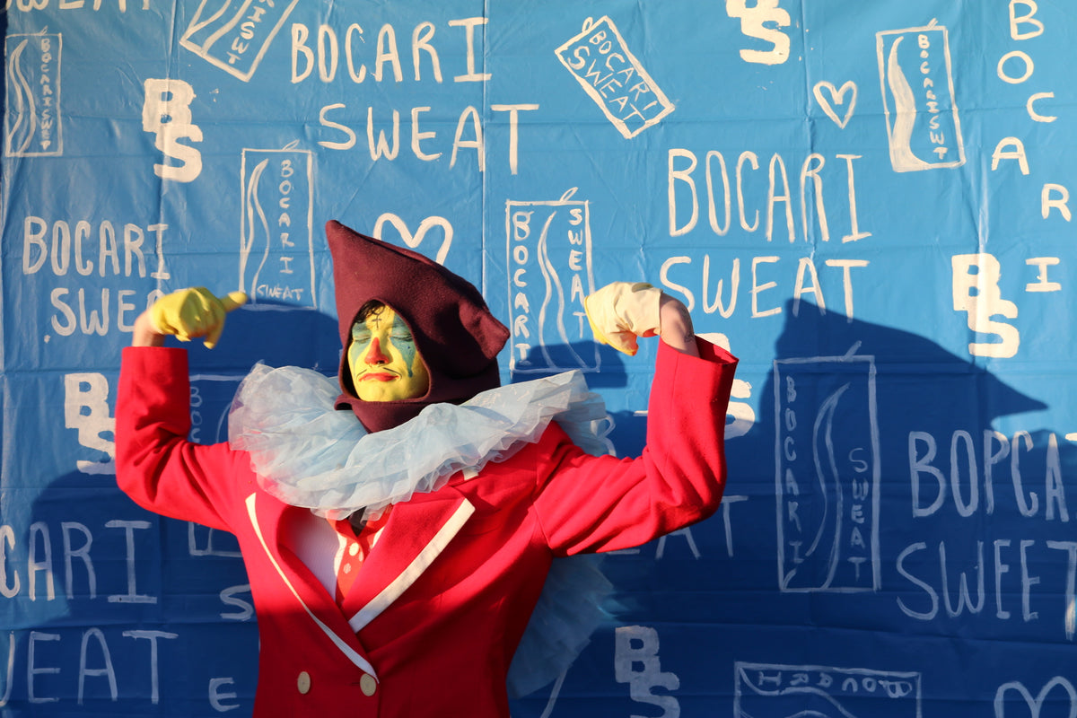 A person in a red costume with a white ruffled collar, yellow gloves, and a dark hat poses with raised arms in front of a blue printed backdrop featuring the words 'BOCARI SWEAT'.
