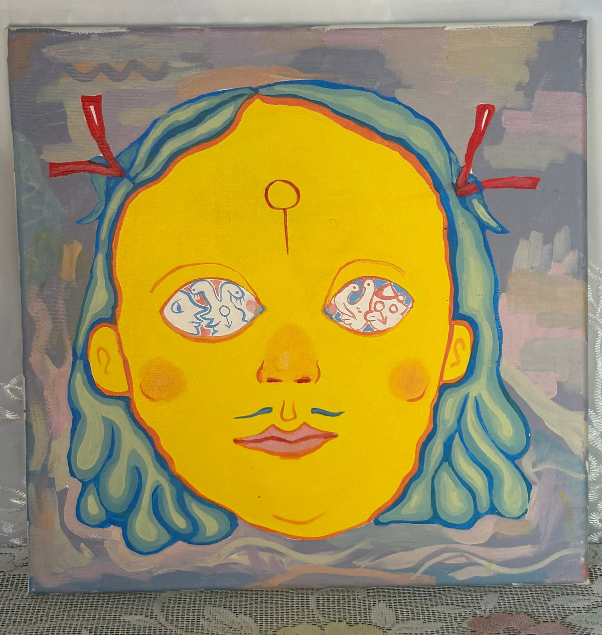 A square canvas painting featuring a stylized yellow face with blue hair and red ribbon accents, leaning against a light gray wall on a lace-covered surface.
