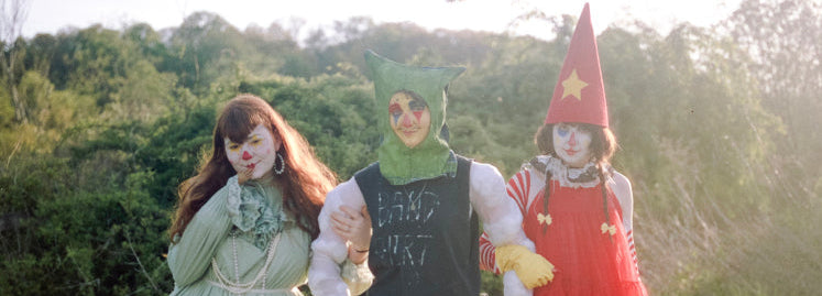Three people standing outdoors on grass wearing elaborate clown-style costumes. The person on the left wears a pale green dress with white patches and white boots.